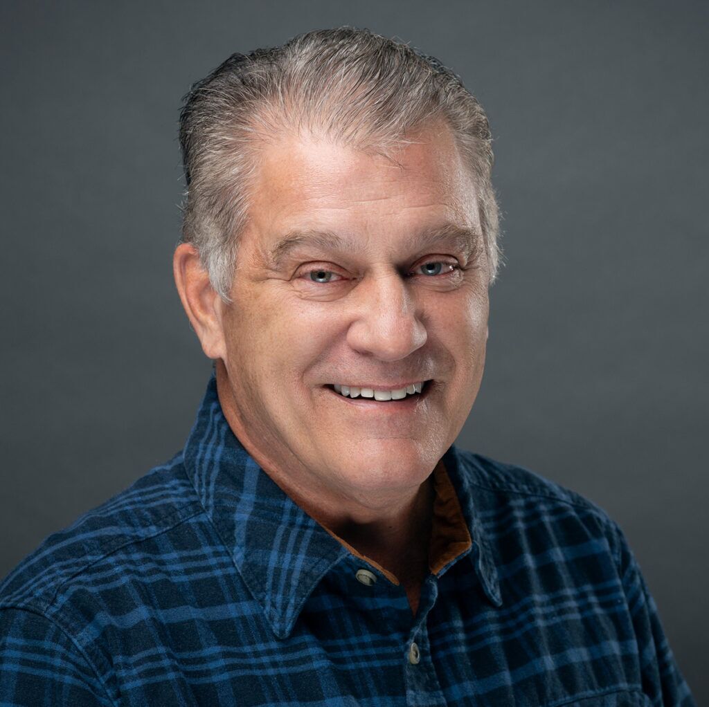 Man with graying hair smiling, wearing a blue plaid shirt against a gray background.